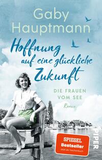 Das Buchcover zeigt eine nostalgische Szenerie mit einem jungen Mädchen in einem weißen Oberteil und grünen Shorts, das lächelnd auf einer Holzbank sitzt. Der Hintergrund ist in sanften Blautönen gehalten und zeigt einen See mit angrenzenden Gebäuden und Vögeln im Flug. Der Titel „Hoffnung auf eine glückliche Zukunft“ ist in geschwungener, blauer Schrift platziert, während der Untertitel „DIE FRAUEN VOM SEE“ in einer klaren, schlichten Schrift steht. Oben sind der Name „Gaby Hauptmann“ und das Label „SPIEGEL Bestseller“ zu sehen.