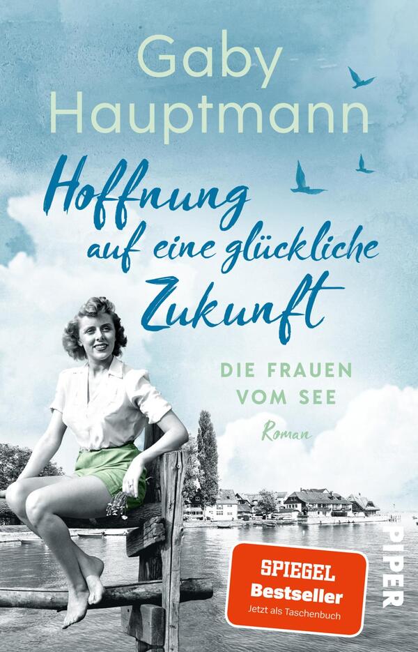 Das Buchcover zeigt eine nostalgische Szenerie mit einem jungen Mädchen in einem weißen Oberteil und grünen Shorts, das lächelnd auf einer Holzbank sitzt. Der Hintergrund ist in sanften Blautönen gehalten und zeigt einen See mit angrenzenden Gebäuden und Vögeln im Flug. Der Titel „Hoffnung auf eine glückliche Zukunft“ ist in geschwungener, blauer Schrift platziert, während der Untertitel „DIE FRAUEN VOM SEE“ in einer klaren, schlichten Schrift steht. Oben sind der Name „Gaby Hauptmann“ und das Label „SPIEGEL Bestseller“ zu sehen.