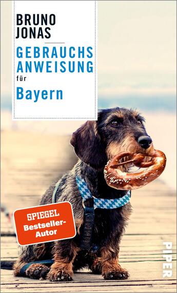 Das Buchcover zeigt einen niedlichen, braunen Dackel mit Schnauze und großen Augen, der auf einem Holzsteg sitzt und eine Brezel im Maul hält. Der Hintergrund ist verschwommen und vermittelt eine entspannte Atmosphäre. Oben links steht in großen, schwarzen Buchstaben „BRUNO JONAS“, darunter in blauer Schrift „GEBRAUCHSANWEISUNG“. Darunter, in kleinerer Schrift, steht „für Bayern“. In der unteren linken Ecke befindet sich ein rotes Quadrat mit weißem Text „SPIEGEL Bestseller-Autor“. Rechts unten ist das Logo des Verlags „Piper“ zu sehen.