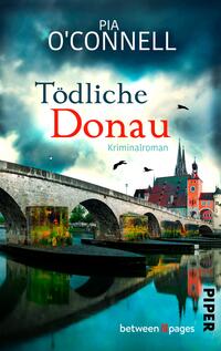 Das Buchcover zeigt eine eindrucksvolle Brücke über die Donau, umgeben von einer malerischen Stadtlandschaft. Der Himmel ist grau und dramatisch, durchzogen von dunklen Wolken. In großen, roten Lettern steht der Titel „Tödliche Donau“ unterhalb von Pia O'Connell, während der Untertitel „Kriminalroman“ in kleineren, weißen Buchstaben zu lesen ist. Silhouetten von Vögeln fliegen über die Szene und tragen zur spannungsgeladenen Atmosphäre bei.