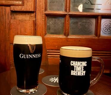 Ein Glas dunkles Guinness und ein Glas mit der Aufschrift „Changing Times Brewery“ stehen nebeneinander auf einem Holztisch. Im Hintergrund sind dekorative Holzrahmen und Fensterscheiben mit Ornamenten sichtbar. 