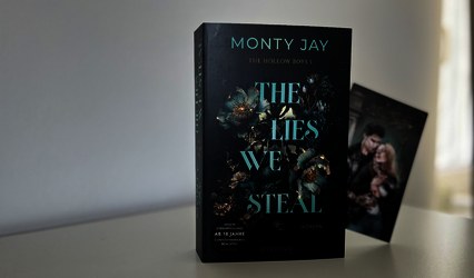 „The Lies we steal“ steht auf einem weißen Tisch vor einer weißen Wand. Aus dem Buch spitzelt die in der ersten Auflage beiliegende Character Card.