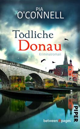 Das Buchcover zeigt eine malerische Brücke, die über einen Fluss führt, unter einem dramatisch bewölkten Himmel. Im Hintergrund sind historische Gebäude mit Türmen zu sehen. Der Titel „Tödliche Donau“ hebt sich in großen, roten Buchstaben hervor, während der Untertitel „Kriminalroman“ in dezentem Blau geschrieben ist. Der Autorname „Pia O‘Connell“ steht oben in eleganter Schrift. Die Farben sind überwiegend kühl und erzeugen eine spannende, mysteriöse Atmosphäre.
