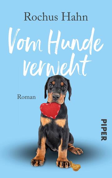 Das Buchcover zeigt einen niedlichen Rottweiler-Welpen, der auf einem hellblauen Hintergrund sitzt. Der Hund hält ein rotes Herzspielzeug im Mund und hat eine verspielte Ausstrahlung. Der Titel „Vom Hunde verweht“ ist in einer freundlichen, handschriftlichen Schriftart in Weiß und gut lesbar platziert. Darunter steht das Wort „Roman“ in einer schlichten Schrift. Unten rechts ist das Logo des Verlags „PIPER“ in Schwarz abgedruckt.