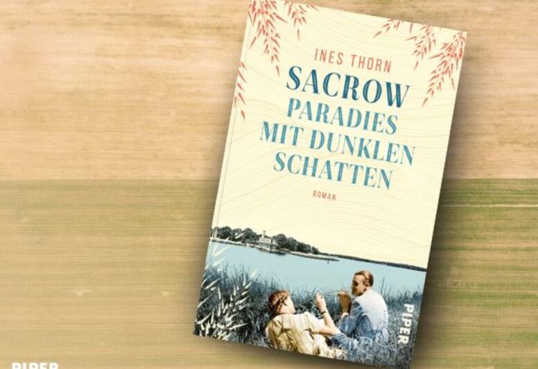 Das Buch 'Sacrow – Paradies mit dunklen Schatten' von Ines Thorn liegt auf einem blauen Holztisch. Daneben steht eine rote Tasse mit Kaffee. Auf dem Cover sind zwei Personen am Ufer des Wannsees gebildet