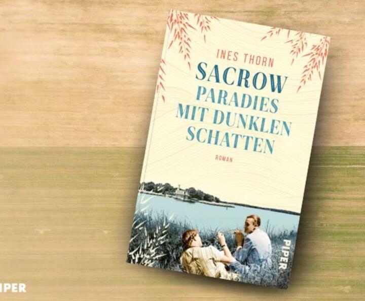 Das Buch 'Sacrow – Paradies mit dunklen Schatten' von Ines Thorn liegt auf einem blauen Holztisch. Daneben steht eine rote Tasse mit Kaffee. Auf dem Cover sind zwei Personen am Ufer des Wannsees gebildet