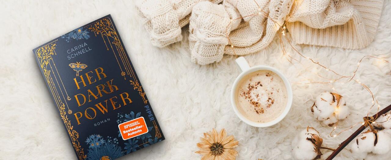 „Her Dark Power“ von Carina Schnell liegt auf einem flauschigen Untergrund. Daneben eine Tasse mit Cappuccino, eine Baumwollast sowie ein gestrickter Pulli.