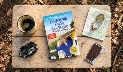 Auf einem Holztisch liegen „Tietjen sucht das Weite“, ein Kompass auf einer Karte, eine Stirnlampe, Tafel Schokolade sowie eine Tasse Kaffee.