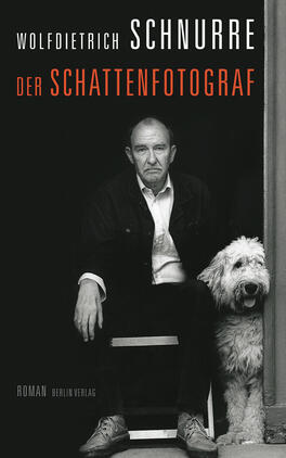 Das Buchcover zeigt einen Mann mittleren Alters mit einem nachdenklichen Ausdruck, der auf einem Stuhl sitzt. Er trägt ein schwarzes Jackett und ein helles Hemd. Neben ihm sitzt ein weißer Hund. Der Hintergrund ist in Grautönen gehalten. Der Titel „DER SCHATTENFOTOGRAF“ ist in großen, roten Buchstaben oben platziert, gefolgt vom Autorennamen „WOLFDIETRICH SCHNURRE“ in Weiß. Am unteren Rand steht das Wort „Roman“ in kleineren Buchstaben. Die gesamte Atmosphäre vermittelt eine melancholische und nachdenkliche Stimmung.