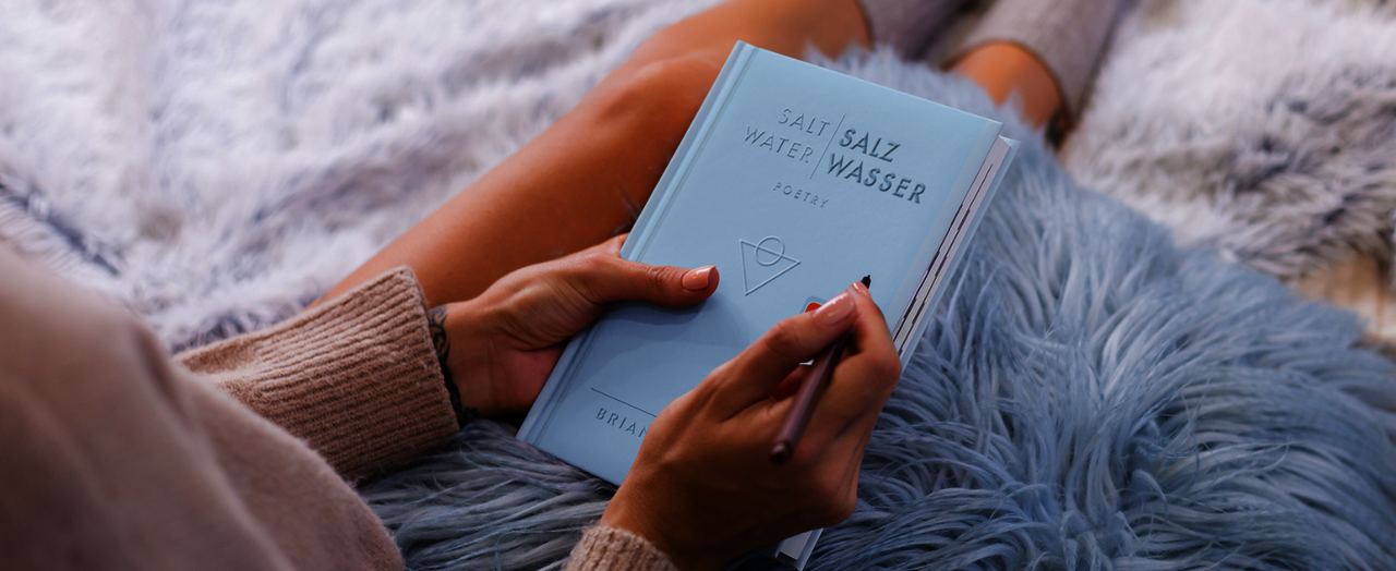 Eine Person in einem beigen Pullover sitzt auf einer weichen, hellblauen Decke. Sie hält „Salt Water / Salz Wasser“ in der Hand.