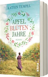 Das Buchcover zeigt eine idyllische Landschaft mit blühenden Apfelbäumen und einer lichtdurchfluteten Wiese. Im Vordergrund steht eine Frau in einem einfachen, lilafarbenen Kleid, die in die Ferne blickt. Über ihr ist der Titel „APFEL BLÜTEN JAHRE“ in einer verspielten, grünen Schriftart platziert, umrahmt von einem Kranz aus bunten Blüten und Äpfeln. Darunter steht das Wort „Roman“ in schlichter, kleiner Schrift. Der Hintergrund ist in sanften Himmelstönen gehalten, wodurch eine verträumte Atmosphäre entsteht. 