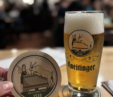 Ein Glas helles Bier steht auf einem Tisch. Daneben wird der Bierdeckel der Brauerei Kneitinger hochgehalten. Darauf sieht man die Zeichnung von einem Mann, der in einem Bier-Trog rührt, aus dem ein Ziegenbock trinkt. Im Hintergrund sind verschwommen die Gäste und Lichter in einem Gastraum zu erkennen.