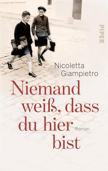 Das Buchcover zeigt ein nostalgisches, schwarz-weißes Foto von zwei Jungen, die in einer Straßenkulisse stehen und miteinander interagieren. Die Jungen tragen klassische Kleidung, einschließlich einer Mütze und kurzen Hosen. Der Titel „Niemand weiß, dass du hier bist“ erscheint in großen, rot gefärbten Buchstaben, während der Name der Autorin, Nicoletta Giampietro, in kleinerer Schrift unterhalb des Titels platziert ist. Am oberen rechten Rand ist das Verlagslogo ‚PIPER‘ sichtbar. Die gesamte Gestaltung vermittelt eine Atmosphäre von Kindheit und Unbeschwertheit.
