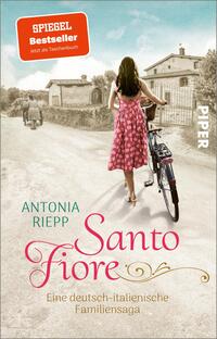 Das Buchcover zeigt eine Wanderin in einem pinken Sommerkleid, die mit einem Fahrrad auf einem ländlichen Weg unterwegs ist. Im Hintergrund sind rustikale Gebäude und grüne Bäume sichtbar, die eine mediterrane Atmosphäre schaffen. Der Titel „Santo Fiore“ ist in geschwungener, roter Schrift platziert, während der Autor „Antonia Riepp“ in einem klaren, blauen Schriftzug darüber steht. Oben rechts befindet sich ein rotes Etikett mit der Aufschrift „SPIEGEL Bestseller – Jetzt als Taschenbuch“. Das Cover strahlt eine familiäre und nostalgische Stimmung aus.
