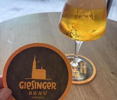 Ein geschwungenes Glas mit hellem Bier und Schaumkrone steht auf einem runden Holztisch. Der Bierdeckel der Giesinger Brauerei wird hochgehalten, darauf sind in Orange der Schriftzug der Brauerei und eine Kirche auf schwarzem Untergrund dargestellt. Im Hintergrund sieht man verschwommen ein paar Holzstühle und Gäste.