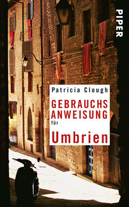 Das Buchcover zeigt eine enge Gasse in einem italienischen Dorf, mit warmen, erdigen Farben und rotem Stoff, der aus Fenstern hängt. Im Vordergrund ist ein schwarzes Rollerfahrzeug zu sehen. Der Titel „GEBRAUCHSANWEISUNG für Umbrien“ ist in großen, weißen und roten Buchstaben zentral positioniert, während der Autorennamen „Patricia Clough“ darüber in schlichter, eleganter Schrift steht. Rechts oben ist der Verlagsname „PIPER“ in weißer Schrift auf schwarzem Hintergrund platziert.