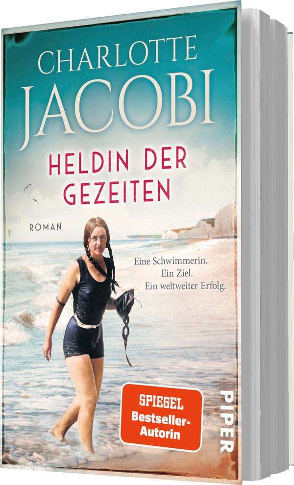 Das Buchcover zeigt eine Frau in einem nostalgischen Schwimmanzug am Strand, die mit einem Schwimmseil in der Hand auf den Ozean blickt. Der Hintergrund ist in sanften Blautönen gehalten, was eine ruhige, aber kraftvolle Atmosphäre vermittelt. Der Titel „Heldin der Gezeiten“ ist auffällig in roten Buchstaben platziert, ergänzt durch die Hinweise „Roman“ und „SPIEGEL Bestseller-Autorin“. Der Name „Charlotte Jacobi“ ist in eleganten, hellen Buchstaben oben auf dem Cover hervorgehoben.