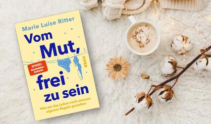 Das Buch „Vom Mut, frei zu sein“ liegt neben einer Tasse Kaffee auf einem weißen flauschigenTeppich