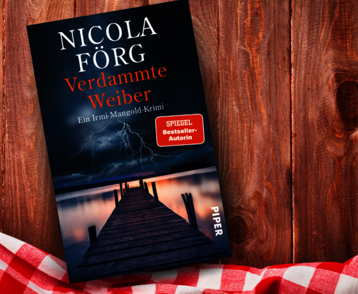 as Buch 'Verdammte Weiber' von Nicola Förg liegt auf einem Holztisch. Auf dem Cover ist ein Steg, der in einen stürmischen Himmel mit Blitzen ragt. Daneben liegt ein rotes kariertes Tuch.