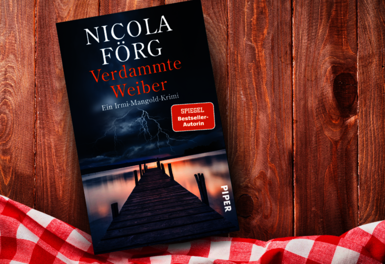 as Buch 'Verdammte Weiber' von Nicola Förg liegt auf einem Holztisch. Auf dem Cover ist ein Steg, der in einen stürmischen Himmel mit Blitzen ragt. Daneben liegt ein rotes kariertes Tuch.