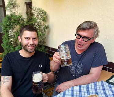 Ein Mann und der Autor sitzen an einem Holztisch mit blau-weißem Tischtuch. Beide halten große Glaskrüge mit dunkelbraunem Bier in der Hand. Auf dem Tisch liegen drei Bierdeckel der Brauerei „Spezial“. 