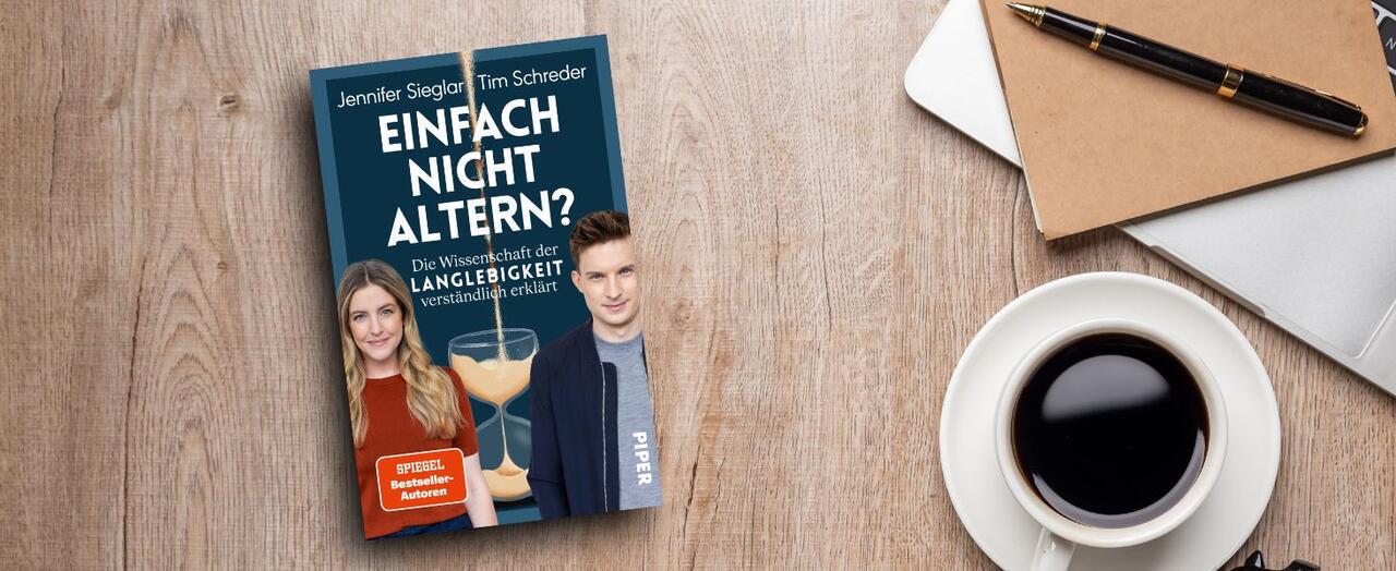 „Einfach nicht altern?“ von Jennifer Sieglar und Tim Schreder liegt neben einer Tasse mit Kaffee, einem Notizbuch und einem Stift auf einem hölzernen Untergrund.