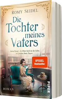 Das Buchcover zeigt eine nostalgische Szene mit einer sitzenden Frau in einem blauen Mantel. Sie hat braune Wellenhaare und blickt nachdenklich auf ein Buch in ihren Händen. Der Hintergrund zeigt eine elegante Bibliothek mit Regalen voller Bücher. Die Farbgebung ist hell und sanft, dominiert von Creme- und Blautönen. Der Titel „Die Tochter meines Vaters“ steht in geschwungener Schrift oberhalb der Frau, während „SPIEGEL Bestseller“ in einem roten Kasten hervorgehoben ist. Der Autorennamen Romy Seidel ist oben vermerkt.