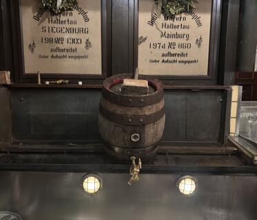 Ein dunkles Holzfass mit goldenem Zapfhahn steht vor einer rustikal getäfelten dunklen Wand. Links und rechts des Fasses hängen Hopfenkränze vor Schildern mit historischen Aufschriften.