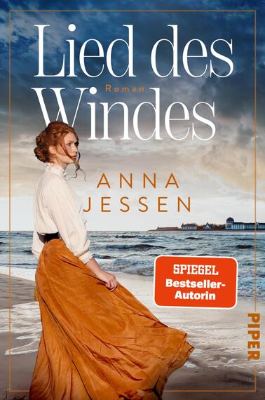 Das Buchcover zeigt eine vom Wind umwehte Frau in historischer Kleidung am Strand, seitlich stehend, rotes Haar hochgesteckt, weiße Bluse und weites ockerfarbenes Rockteil. Im Hintergrund stürmisches Meer und wolkiger Himmel, seitlich ein Pier. Der Titel „Lied des Windes“ ist groß in weißen Serifbuchstaben, darunter der Autorenname ANNA JESSEN in orangeen Großbuchstaben. Ein orange-roter Button mit dem Text „SPIEGEL Bestseller-Autorin“.