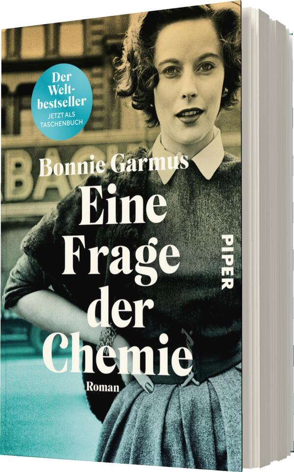 Das Buchcover zeigt eine nostalgische Schwarz-Weiß-Fotografie einer Frau mit lockigem Haar, die direkt in die Kamera blickt. Sie trägt ein elegantes, schlichtes Outfit und hat eine selbstbewusste Ausstrahlung. Der Titel „Eine Frage der Chemie“ steht in großer, weißer Schrift im unteren Drittel des Covers, während der Name der Autorin, Bonnie Garmus, darüber in kleinerer Schrift platziert ist. Ein blauer Button links oben verkündet „Der Welterfolg – Jetzt als Taschenbuch“. Das Cover vermittelt eine ansprechende und inspirierende Atmosphäre.