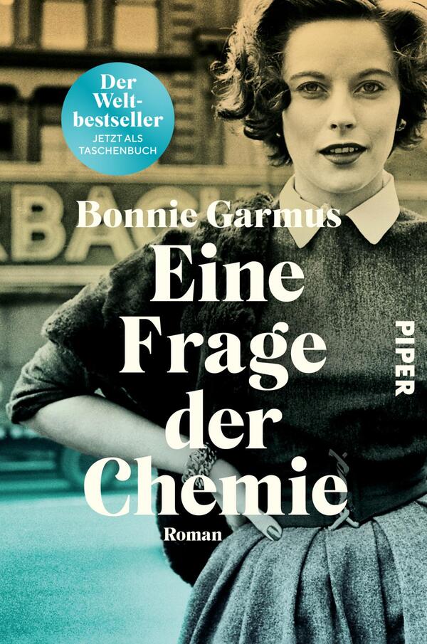 Das Buchcover zeigt eine schwarz-weiß gehaltene Fotografie einer Frau in einem eleganten grauen Kleid mit weißem Kragen. Sie hat lockige Haare und blickt direkt in die Kamera. Im Hintergrund ist ein unscharfer Schriftzug zu erkennen. Oberhalb der Frau steht in kräftigem Weiß der Titel „Eine Frage der Chemie“, gefolgt vom Zusatz „Roman“ in kleinerer Schrift. Darunter ist der Name der Autorin, Bonnie Garmus, platziert. Oben links befindet sich ein blauer Kreis mit dem Text „Der Bestseller“ und „Jetzt als Taschenbuch“.