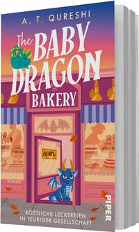 Das Buchcover zeigt eine verspielte, rosa-violette Bäckerei-Fassade vor einem orange-violetten Himmel. Groß oben steht der Autorenname A. T. Qureshi in Gelb, der Titel „The Baby Dragon Bakery“ in großer weißer, verspielter Serifschrift. Im Mittelpunkt sitzt ein blauer Babydrache in der Tür; im Fenster sind Kuchen und warme Lampen zu sehen. Ein kleines Schild im Türfenster zeigt „open“. Unter dem Bild steht der Untertitel „Köstliche Leckereien in feuriger Gesellschaft“.