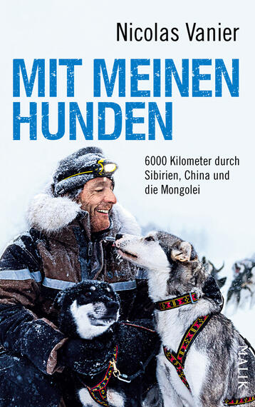 Das Buchcover zeigt einen grinsenden Mann mit dunklem Bart in einer winterlichen Landschaft, umgeben von mehreren Hunden. Die Tiere sind meist in Grau- und Schwarztönen gehalten, während der Mann eine warme, helle Jacke trägt. Der Titel „MIT MEINEN HUNDEN“ ist in großen, hellblauen Buchstaben prominent platziert, unter dem sich der Untertitel „6000 Kilometer durch Sibirien, China und die Mongolei“ in schlichter Schrift befindet. Der Autorname „Nicolas Vanier“ steht oben auf dem Cover in dezentem Schwarz.
