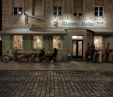 Das Gebäude der Brauerei „Manns Bräu“ ist bei Nacht beleuchtet. Über dem Eingang hängt ein Schild mit dem Brauereinamen, davor sitzen und stehen Leute an Holztischen. Das Kopfsteinpflaster reflektiert das Licht. 