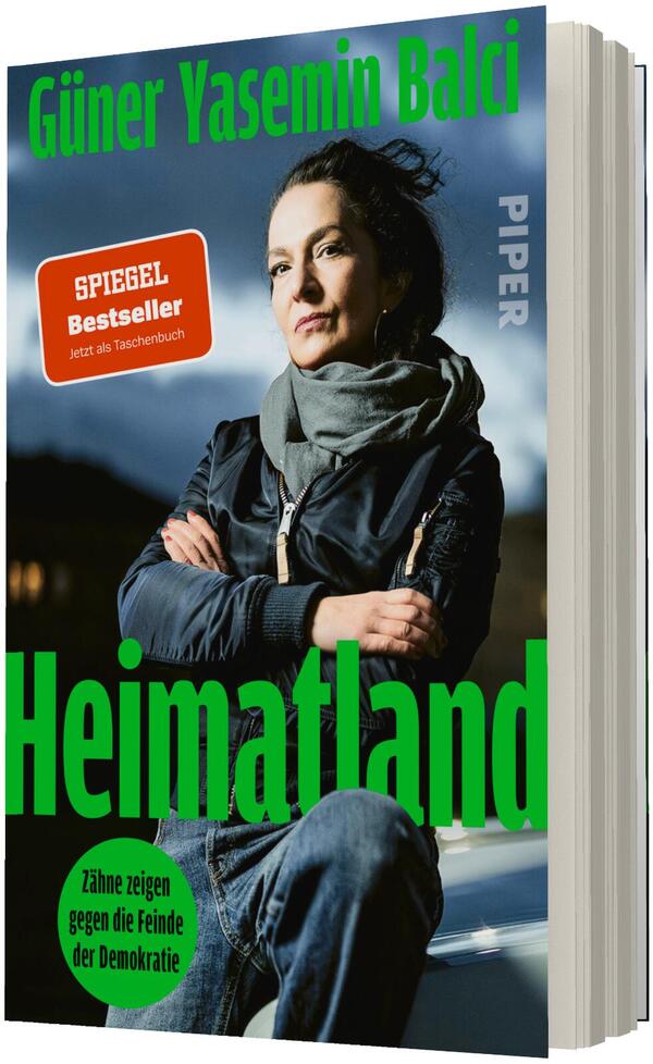 Das Buchcover zeigt eine Porträtfotografie der Autorin Güner Yasemin Balci, die mit verschränkten Armen, dunkler Jacke und grauem Schal nachdenklich in die Ferne blickt; dunkler, wolkiger Himmel im Hintergrund. Großflächiger, kräftiger grüner Titel „Heimatland“ unten, Autorenname oben ebenfalls grün. Oranger quadratischer Sticker „SPIEGEL Bestseller Jetzt als Taschenbuch“ links oben und kleiner grüner Kreis mit Untertitel „Zähne zeigen gegen die Feinde der Demokratie“. Verlagslogo nicht beschrieben.