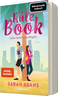 Das Buchcover zeigt einen sonnigen Strand mit Palmen, in sanften Pastellfarben. Im Vordergrund stehen zwei Figuren: ein Mann in sportlicher Kleidung mit der Nummer 82 und eine Frau in einem pinken Kleid, die zusammen lachen. Über dem Bild ist der Titel „The Rule Book“ in großen, auffälligen Buchstaben zu sehen, gefolgt von „Liebe kennt keine Regeln“ in kleinerer Schrift. Unten steht der Name der Autorin „SARAH ADAMS“ und das Wort „ROMAN“. In der oberen Ecke befindet sich ein Hashtag.