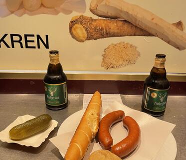 Zwei gedrungene dunkle Flaschen Hubertus-Bier stehen neben einem Teller mit Würstchen, Senf und der für Wien typischen Salzstangerl, einem länglichen Weißgebäck mit Salz und Kümmel. Auf einem separaten Pappteller liegt eine Gewürzgurke. An der Wand dahinter sind Bilder von Zutaten wie Silberzwiebeln und Meerrettich angebracht.