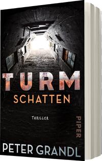 Das Buchcover zeigt einen düsteren tunnelartigen Raum mit einem hellen Lichtstrahl am Ende, der eine übersinnliche Atmosphäre erzeugt. Die Wände sind aus grauem Stein, und Fensteröffnungen sind erkennbar. Die Titelschrift „TURM SCHATTEN“ ist groß und in einem kontrastierenden, leicht verwitterten Rot, während „THRILLER“ in einfacher weißer Schrift darunter steht. Der Autorennamen „PETER GRANDL“ erscheint in heller, minimalistischer Typografie am unteren Rand des Covers.