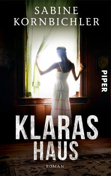 Das Buchcover zeigt eine Frau in einem weißen Kleid, die an einem Fenster steht und nach draußen blickt. Der Raum ist in dunklen Tönen gehalten, was eine geheimnisvolle Atmosphäre erzeugt. Helle Lichtstrahlen fallen durch die Vorhänge und beleuchten die Figur. Der Titel „KLARAS HAUS“ wird in großen, weißen Buchstaben dargestellt, während der Autorenname „SABINE KORNBICHLER“ oben in einer eleganten Schriftart steht. Am rechten Rand ist das Logo des Verlags „PIPER“ sichtbar.