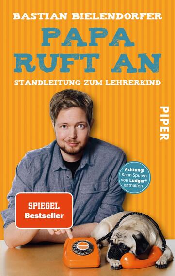 Das Buchcover zeigt einen lächelnden Mann mit kurzes, helles Haar, der einen blauen Pullover trägt. Er sitzt vor einem orangefarbenen Hintergrund und hält ein typisches Telefon in der Hand. Unter dem Titel „PAPA RUFT AN“ steht der Untertitel „STANDLEITUNG ZUM LEHRERKIND“ in großen, hellblauen Buchstaben. Der Titel und der Untertitel sind klar und auffällig gestaltet. In der oberen linken Ecke prangt der Hinweis „SPIEGEL Bestseller“. Am oberen Rand ist ein Warnhinweis angebracht: „Achtung! Kann Spuren von Ludger enthalten.“ Das Verlagslogo „PIPER“ befindet sich unten rechts.