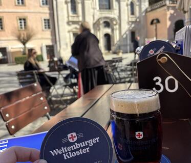 Ein Glas mit dunkelbraunem Bier steht auf einem Tisch im Hof des Weltenburger Klosters. Ein dunkelblauer Bierdeckel mit Klosterlogo wird hochgehalten. Im Hintergrund sind verschwommen die Klosterfassade und ein paar Menschen zu sehen.