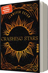 Das Buchcover zeigt einen dunklen, fast schwarzen Hintergrund mit einem glitzernden Sternenhimmel. In der Mitte befindet sich eine große, goldene Sonne mit geschwungenen Strahlen, die dynamisch und feurig wirken. Oberhalb der Sonne steht in goldener Schrift der Name „Jennifer Estep“. Darunter ist der Titel „CRASHING STARS“ in großem, auffälligem Schriftzug platziert. Unten rechts befindet sich das Wort „PIPER“ in kleinerer Schrift. Ein rotes Banner spricht von der Autorin als „SPIEGEL Bestsellerautorin“.