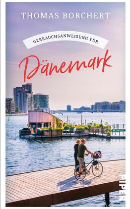 Das Buchcover zeigt eine idyllische Küstenlandschaft mit sanften Wellen und modernen Gebäuden im Hintergrund. Im Vordergrund steht ein Paar auf einer Holzplanke, wobei eine Frau auf einem Fahrrad sitzt. Der Titel „Gebrauchsanweisung für Dänemark“ ist in einer auffälligen roten Schriftart gehalten, während der Autor „Thomas Borchert“ oben in einer klassischen Schriftart in Weiß präsentiert wird. Der Himmel strahlt in sanften Pastelltönen, was eine entspannte Atmosphäre vermittelt.