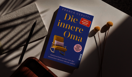 „Die innere Oma“ von Franca Cerutti liegt schräg auf einem hellen Tisch. Daneben liegen ein graues Notizbuch, ein schwarzer Stift und getrocknete Blumen.