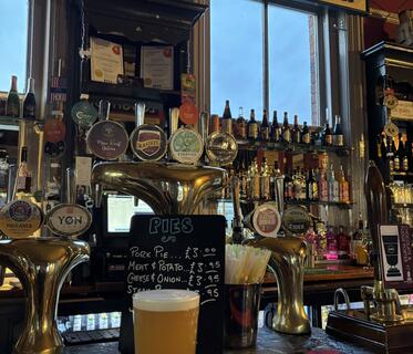 Ein Glas helles Bier steht auf einer Theke. Ein Schriftzug an der Wand lautet „Stalybridge Station“. Darunter sind goldene Zapfhähne, ein Schild mit Pies-Angeboten und in Regalen an den Fenstern Spirituosen sichtbar.