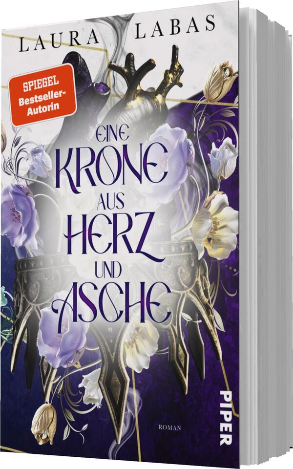 Das Buchcover zeigt eine stilisierte Krone aus goldenen und schwarzen Elementen, umgeben von blühenden lila und weißen Blumen. Der Titel „EINE KRONE AUS HERZ UND ASCHE“ ist in großen, geschwungenen, goldenen Buchstaben in der Mitte platziert. Oben steht der Name der Autorin „Laura Labas“ in einer schlichten Schrift. Ein rotes Etikett mit dem Text „SPIEGEL-Bestseller-Autorin“ hebt die Bekanntheit der Autorin hervor. Der Hintergrund ist in einem sanften Violett gehalten, was eine mystische Atmosphäre schafft.