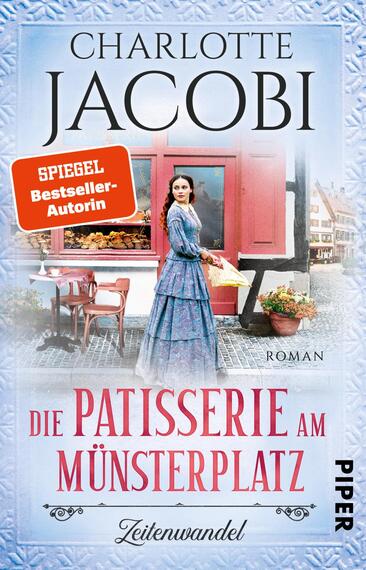 Das Buchcover zeigt eine elegante Frau in einem historischen Kleid, die in einer Patisserie steht und eine Torte in den Händen hält. Der Hintergrund zeigt ein typisch deutsches Stadtbild mit einem roten Fenster und Bäckereiwaren. Der Titel „Die Patisserie am Münsterplatz“ ist in geschwungener Schrift in Rot und Weiß unter dem Bild platziert. Darüber steht „Charlotte Jacobi“ in großen, eleganten Buchstaben. In der oberen linken Ecke befindet sich ein rotes Kästchen mit der Aufschrift „SPIEGEL Bestseller-Autorin“. Die Atmosphäre ist nostalgisch und einladend.
