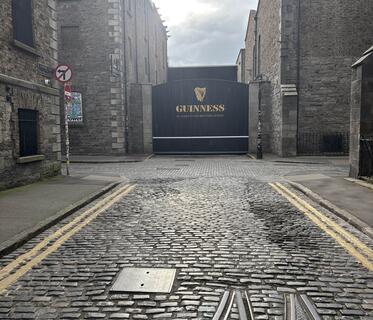 Das schwarze Eingangstor der Guinness-Brauerei in Dublin zeigt das Logo der Brauerei und die Aufschrift „St. James’s Gate Brewery, Dublin“. Im Vordergrund führt eine Kopfsteinpflasterstraße mit alten Schienenfragmenten zum Tor.