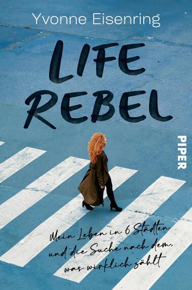 Das Buchcover zeigt eine Frau mit lockigem, orangefarbenem Haar, die auf einem blauen, mit weißen Zebrastreifen versehenen Boden steht. Sie trägt einen dunklen Mantel und eine helle Hose. Der Titel „LIFE REBEL“ ist in großen, fette, schwarzen Buchstaben platziert, während der Untertitel „Mein Leben in 6 Städten und die Suche nach dem, was wirklich zählt“ in einer handschriftlichen Schriftart in Schwarz darunter steht. Das Verlagslogo „PIPER“ ist in kleineren, weißen Buchstaben in der unteren rechten Ecke vorhanden.