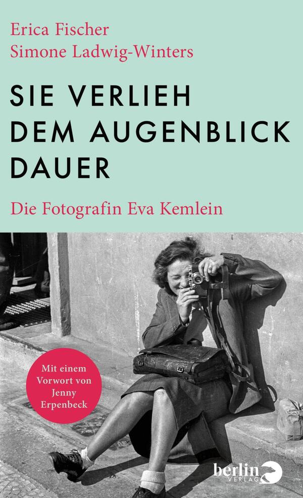 Das Buchcover zeigt im oberen Drittel ein mintgrünes Feld mit dem großen, schwarzen, serifenlosen Titel in Versalien „SIE VERLIEH DEM AUGENBLICK DAUER“; links oben stehen in roter Serifenschrift die Autorennamen Erica Fischer und Simone Ladwig-Winters, darunter in kleiner roter Serifenschrift der Untertitel Die Fotografin Eva Kemlein. Die untere Hälfte zeigt ein schwarzweißes Foto einer lächelnden Frau, die mit einer Kamera auf dem Gehsteig sitzt; Atmosphäre: nostalgisch, dokumentarisch.